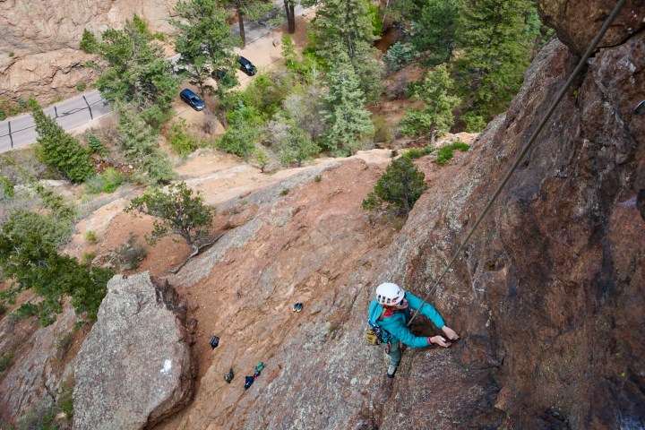 Climber in blue jacket scaling a steep rock face with trees and parked cars below.