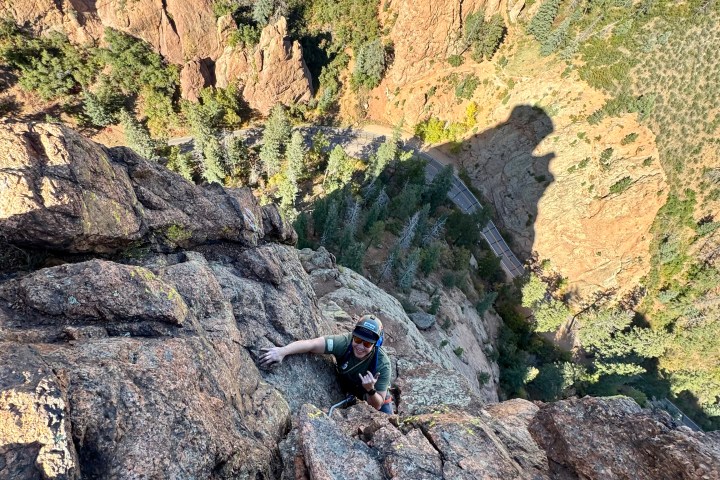 Person climbing steep rocky cliff with trees below, large shadow on rock face.