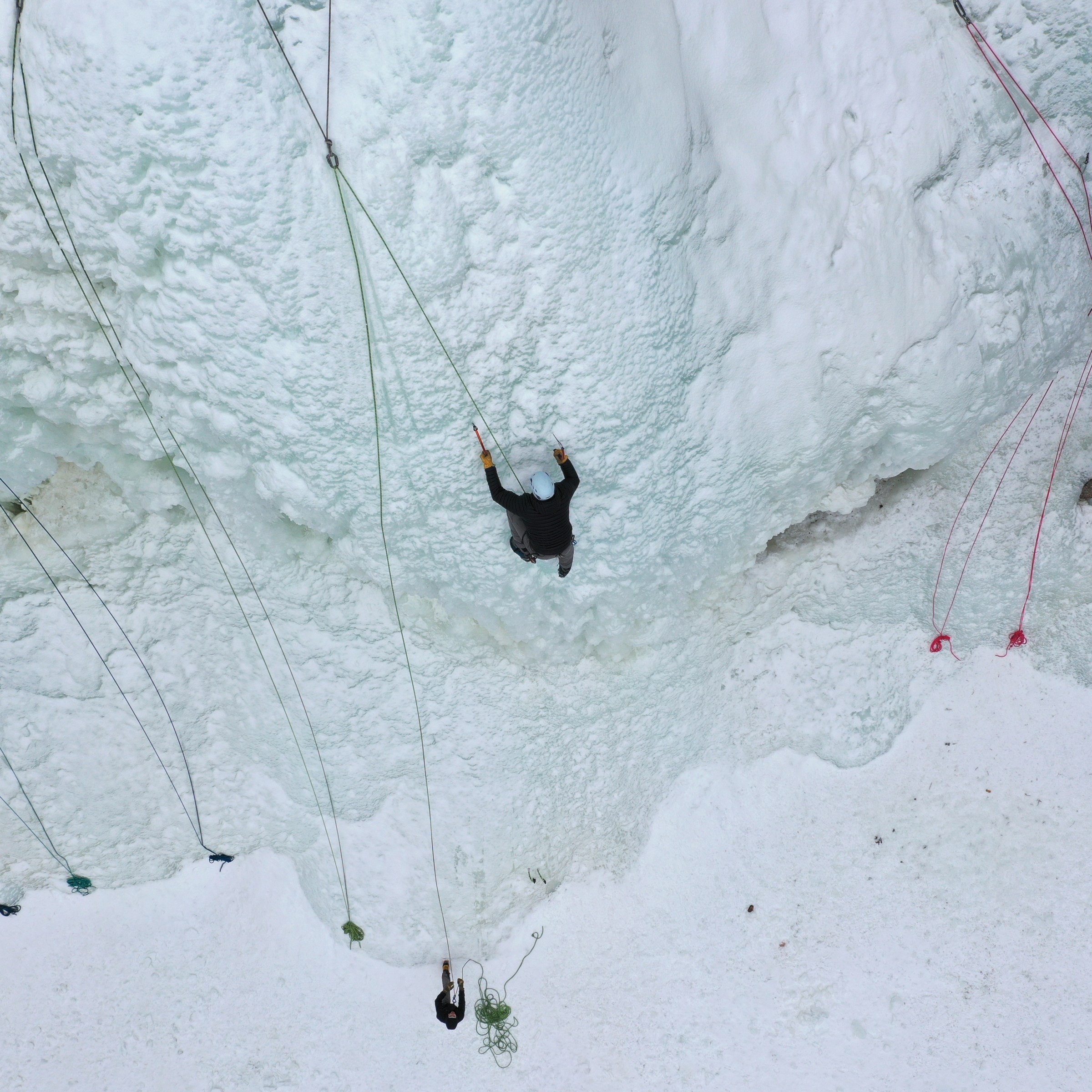 Person ice climbing on a frozen wall with safety ropes, viewed from above.