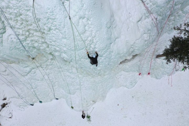Person ice climbing on a frozen wall with safety ropes, viewed from above.