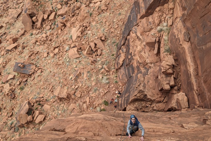 Climber ascending a red rock cliff with a safety rope in a rugged, arid landscape.