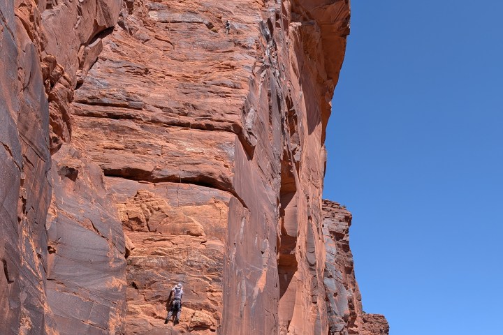 Person rock climbing a tall red sandstone cliff under a clear blue sky.