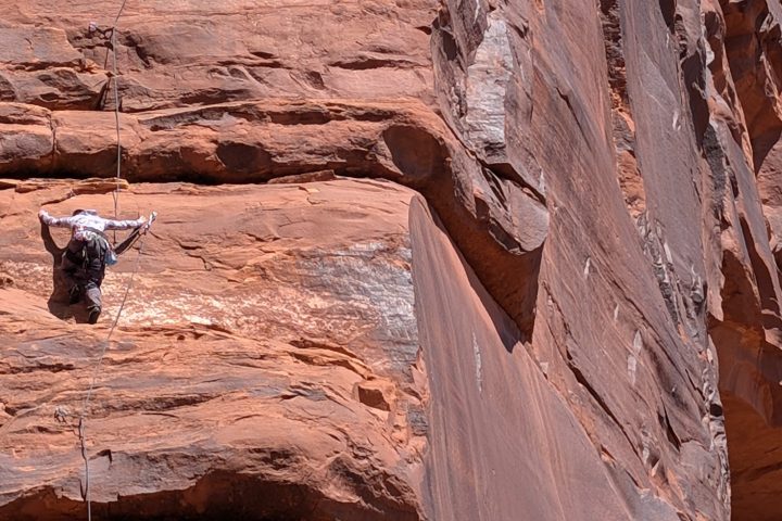Two rock climbers ascend a vertical red rock cliff with ropes.