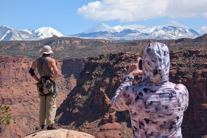 Two people on a cliff edge with snow-capped mountains in the background, one taking a photo.