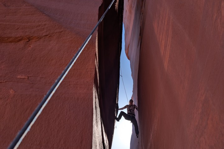 Person rock climbing in a narrow canyon with a rope, surrounded by red sandstone walls.