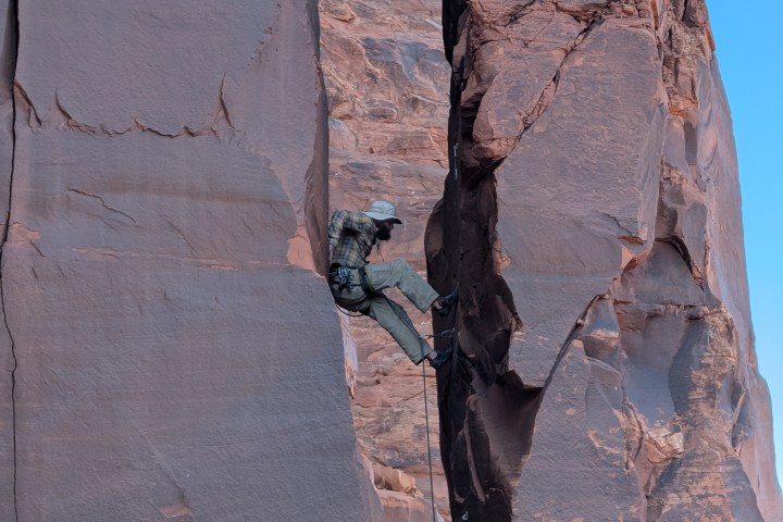 Rock climber ascending a sandstone cliff with safety gear against a clear blue sky.