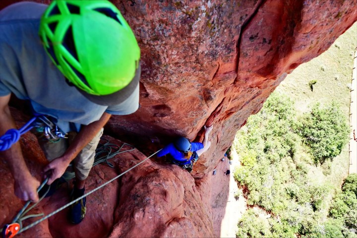 Two climbers scaling a red rock cliff with safety gear and ropes.
