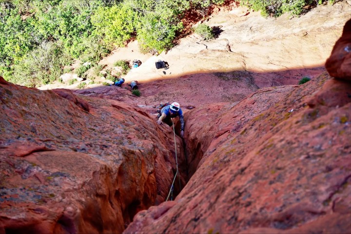 Climber scaling a steep red rock crevice with gear visible above.
