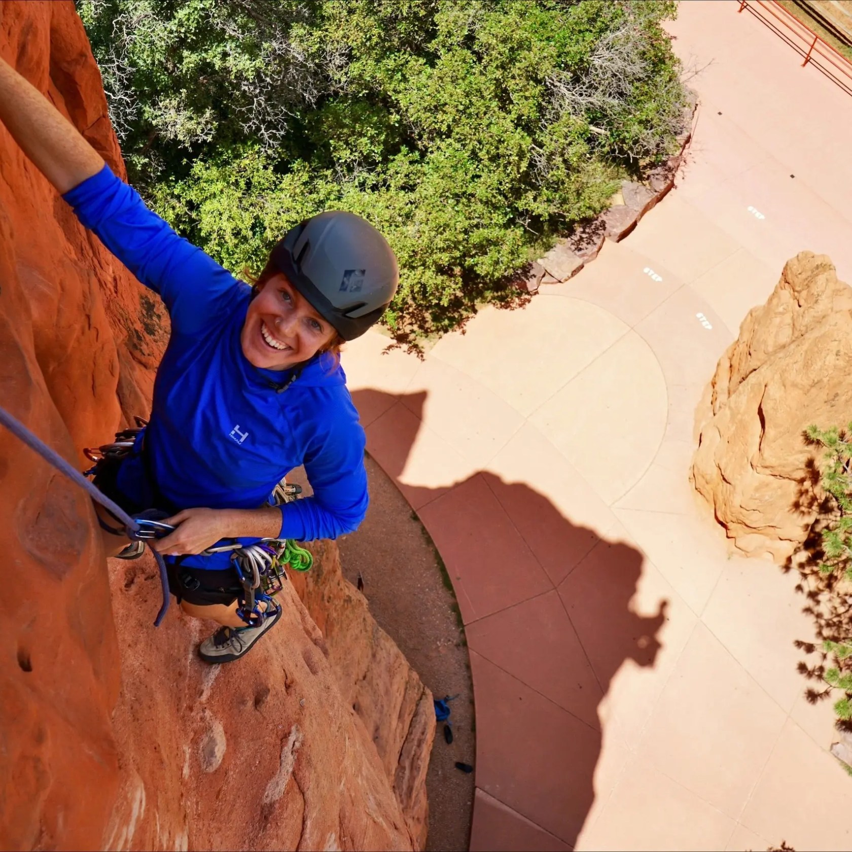 Person rock climbing on a red rock face, smiling, with trees and path below.