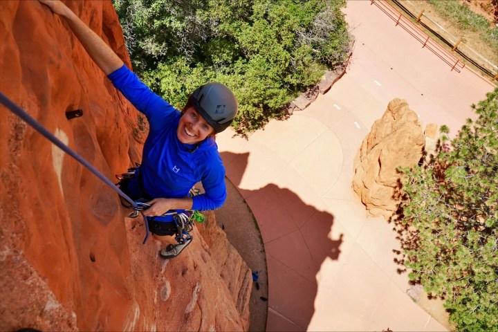 Person rock climbing on a red rock face, smiling, with trees and path below.