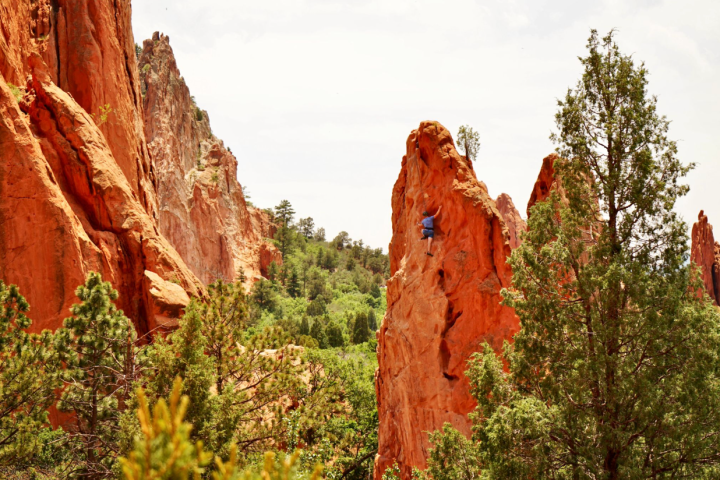 Person rock climbing on tall, red rock formation with trees around, cloudy sky.